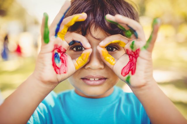 Boy with painted hands making glasses gesture in a park. Perfect for concepts of childhood creativity, outdoor activities, and playful imagination. Suitable for educational materials, children's activity promotions, and family-oriented advertisements.