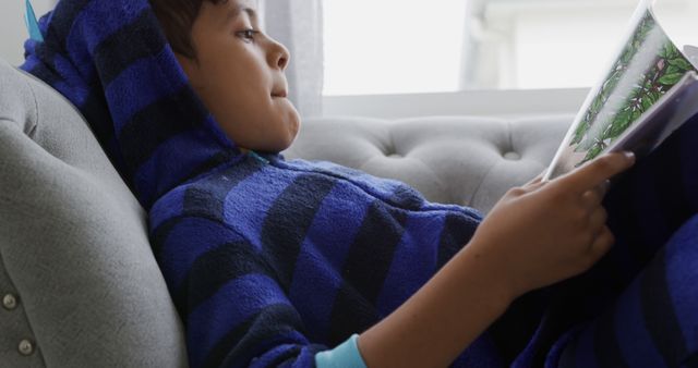 Young boy relaxing on a couch and reading a book while wearing a blue striped monster onesie. This image can be used to depict childhood leisure activities, cozy home settings, and the educational benefits of reading. Ideal for promotions related to children's books, educational content, home comfort products, or parenting resources.