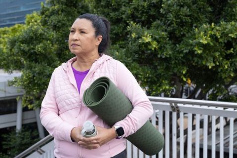 Mature woman pausing before outdoor yoga holding green mat and water bottle wearing pink jacket