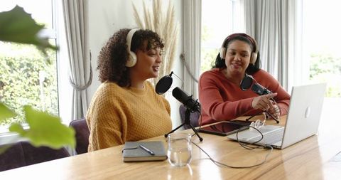 Diverse women cohosting cozy home podcast recording with headphones, microphones, laptop