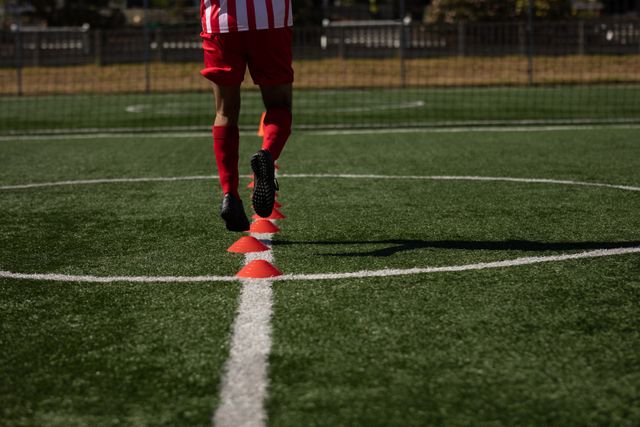 Low section of a football player in a team uniform running between cones on a sports field. Ideal for use in articles about sports training, fitness routines, soccer drills, athletic practice, and team sports. Perfect for illustrating concepts of agility, coordination, and outdoor exercise.