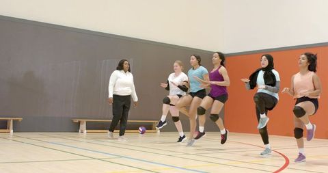 Diverse female volleyball team performing high-knee warm-up drills with coach observing