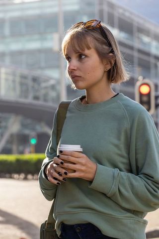 Young female commuter standing on urban sidewalk holding takeaway coffee in green sweatshirt