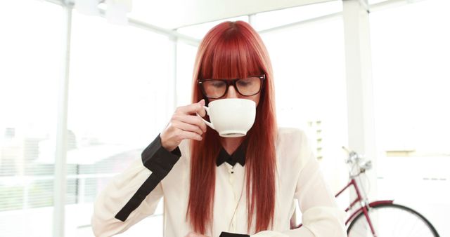 A Caucasian woman with red hair is sipping coffee from a white cup, with copy space. Her attire suggests she might be a professional or businesswoman taking a break in a bright, modern setting.