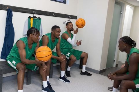 Diverse male basketball team sitting on locker bench spinning ball in green uniforms