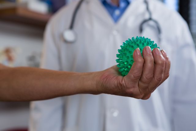 Woman using a green stress ball for hand exercise in a clinical setting. A doctor is visible in the background, suggesting a medical or therapeutic environment. Useful for illustrating concepts of physical therapy, rehabilitation, healthcare, and wellness. Ideal for medical websites, health blogs, and educational materials on hand exercises and recovery.