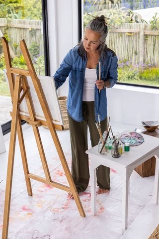 Senior Woman Painting at Easel in Bright Home Studio with Palette, Brushes and Garden View