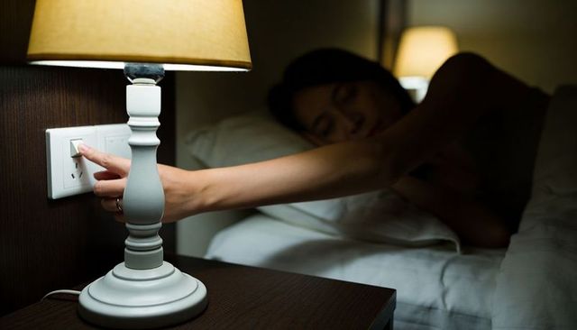 Woman reaching for bedside lamp switch at night in cozy low light bedroom, sleeping under duvet