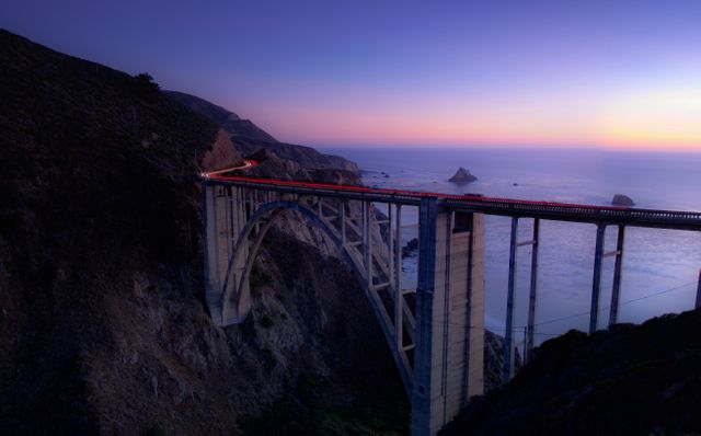 Bixby Creek Bridge stretching over dramatic cliffs with a view of the Pacific Ocean during dusk, creating a picturesque and tranquil scene with coastal beauty and architectural magnificence. Useful for travel ads, tourism promotions, scenic wallpapers, inspirational posters, or websites about landmarks and adventure destinations.