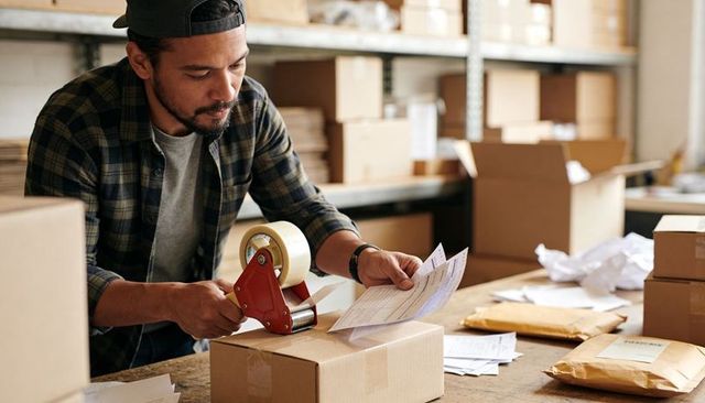 Packing orders in busy warehouse sealing cardboard box with tape dispenser at workstation