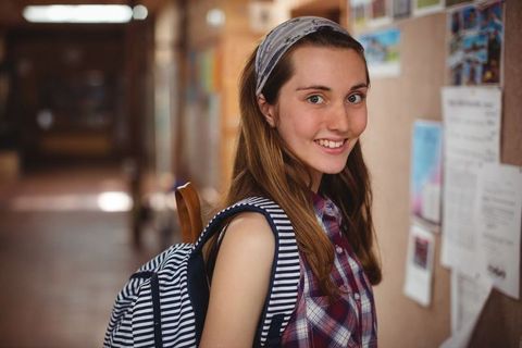 Cheerful teenage girl standing with backpack in school hallway