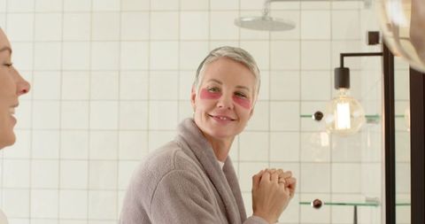 Senior woman and mid-adult companion wearing under-eye gel patches smiling by vanity