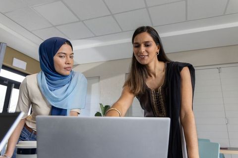 Diverse female colleagues mentoring and collaborating on laptop in modern coworking workspace