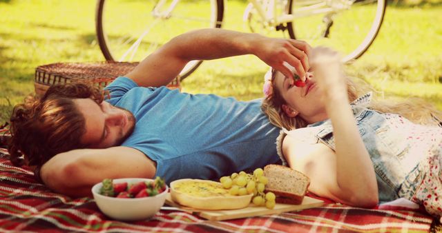 A young Caucasian couple enjoys a romantic picnic in the park, with the woman playfully feeding the man a strawberry, with copy space. Their relaxed posture and the spread of food create a cozy and intimate atmosphere.