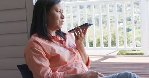 Mature woman speaking into smartphone on sunlit porch, relaxed outdoor conversation