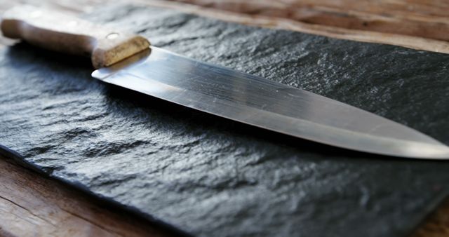 High-quality chef's knife with stainless steel blade and wooden handle lying on textured slate stone surface. Ideal for use in articles, blogs, or advertisements focusing on cooking, culinary skills, kitchen tools, and chef equipment. Also suitable for educational materials on knife handling and care.