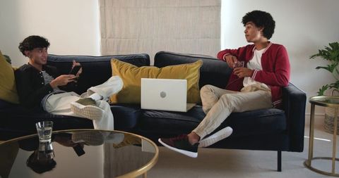 Two young men relaxing on navy sofa using laptop and phone while chatting in living room