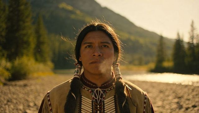 Standing native american man wearing traditional regalia and beaded breastplate by mountain river