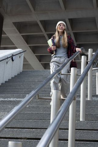 Female student walking down concrete campus stairs holding notebooks wearing beanie
