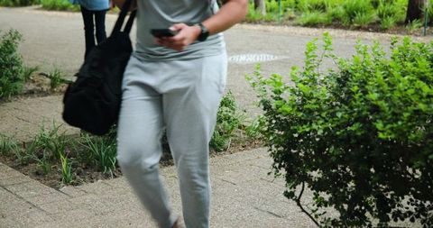 Urban commuter walking with duffel bag while checking phone on leafy campus path