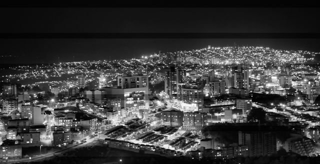 Panoramic black and white view of a bustling cityscape illuminated by numerous city lights at night. Skyscrapers and buildings are prominently featured against a hilly landscape, creating a dramatic and dynamic atmosphere. Perfect for urban development presentations, architectural project backgrounds, and nighttime city-focused publications.