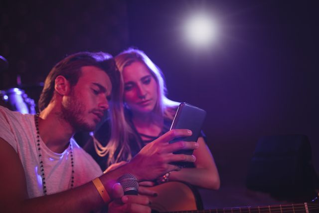 Male and female musicians are using a mobile phone while practicing in a nightclub. The scene is illuminated with stage lights, creating a vibrant atmosphere. This image can be used for themes related to music, technology in entertainment, collaboration, and nightlife events.