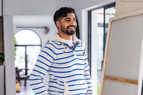 Smiling artist wearing blue striped hoodie standing by canvas in modern minimalist home studio