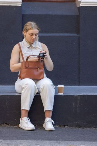 Young woman checking smartphone while sitting on city step with leather handbag and coffee