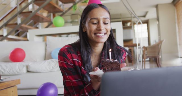 Biracial woman using laptop having birthday image chat holding a cake blowing candle. self isolation during covid 19 coronavirus pandemic.