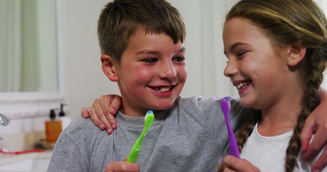 Young brother and sister are brushing their teeth together in a bathroom, smiling and enjoying the moment. They appear to be happy and close, promoting good dental hygiene with their colorful toothbrushes. This is ideal for use in healthcare promotion, dental clinic displays, advertisements focusing on family health, or personal care product advertising.
