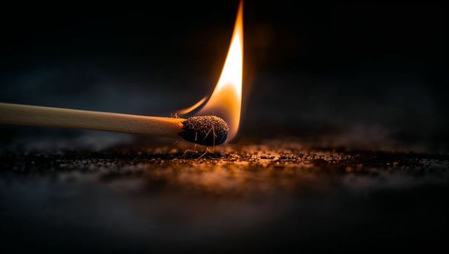 Burning matchstick head glowing with flame and embers on dark textured surface
