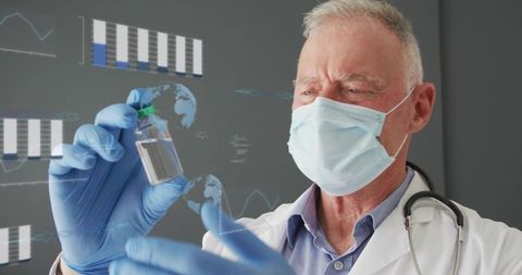 Senior doctor examining vaccine vial while wearing mask and blue gloves with stethoscope