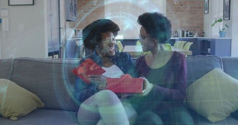 Mother and daughter sharing red gift on sofa with holographic digital overlay for family celebration
