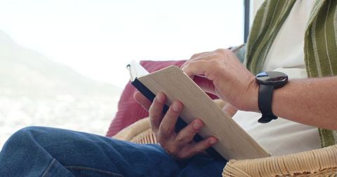 Senior man sitting in wicker armchair writing in journal near sunny window wearing smartwatch
