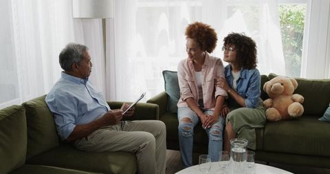 Senior man discussing paperwork with adult daughters on green sofa during family meeting