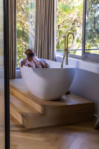 Mature african man relaxing in freestanding bathtub with headphones and gold faucet by window