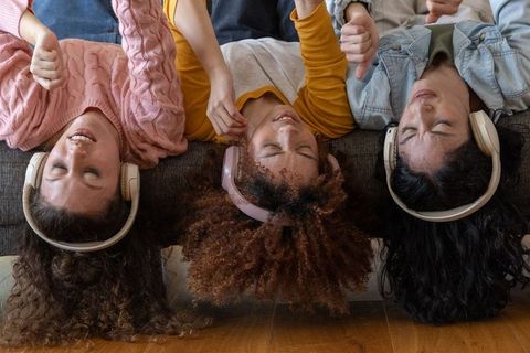 Three friends upside down on sofa wearing headphones, listening to music and laughing