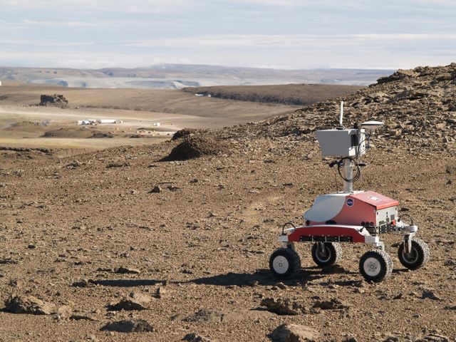 Image of the K-10 rover named 'Red' descending a hill towards the base camp at Haughton Crater on Devon Island, Nunavut, in the Canadian high arctic. The site is part of the NASA-led Haughton-Mars Project, studying the crater and surroundings as Mars analog since 1997. Useful for articles on space exploration, planetary science, and field research on Mars analogs.