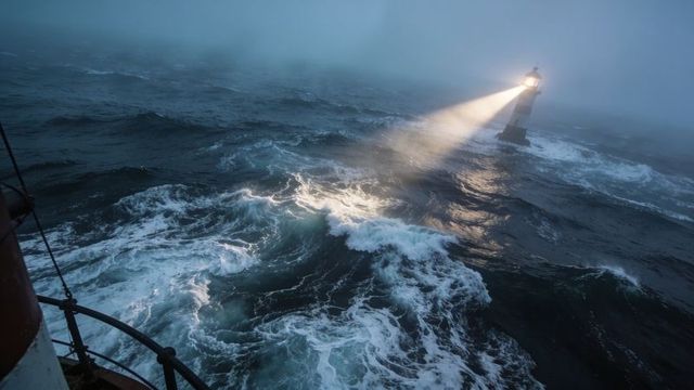 Lighthouse beam cutting through foggy stormy sea with towering waves and ship deck railing