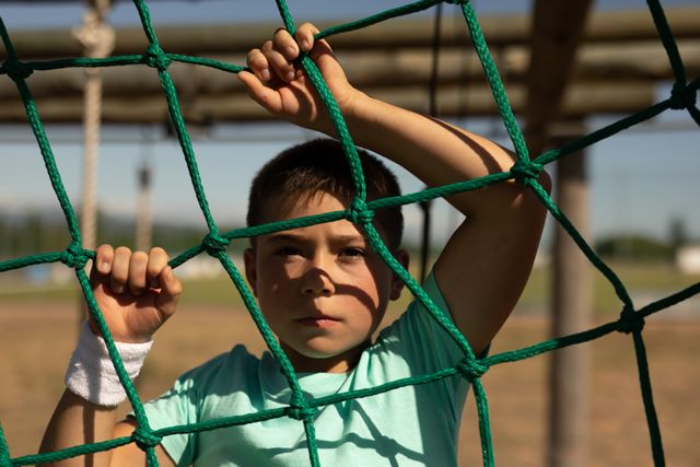 Young boy participating in a boot camp, holding onto green ropes of a jungle gym. Ideal for use in advertisements for children's fitness programs, outdoor activities, summer camps, or educational materials promoting physical health and exercise.
