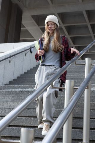 Female student walking down concrete campus stairs holding notebooks touching steel rail