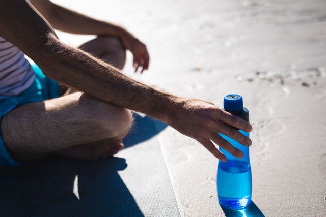 Man sitting cross-legged on a mat, holding a blue water bottle on a sunny day. Ideal for promoting healthy lifestyle, hydration, outdoor activities, fitness routines, and summer relaxation. Suitable for use in health and wellness blogs, fitness websites, and advertisements for outdoor gear.