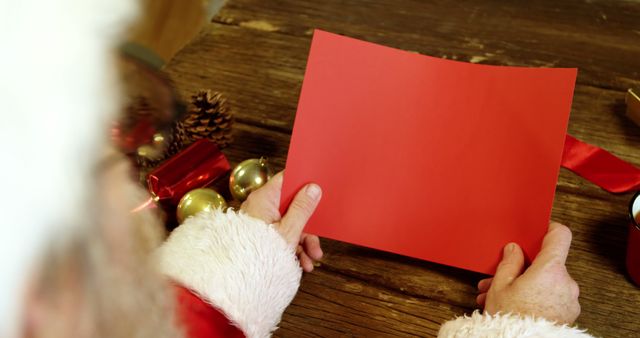 Santa Claus in traditional costume holding blank red paper at wooden table. Surrounded by festive Christmas decorations such as gold ornaments, pine cones, and red ribbons, creating a cozy holiday atmosphere. Ideal for Christmas event promotions, seasonal marketing, holiday greeting card templates, or festive advertisements.