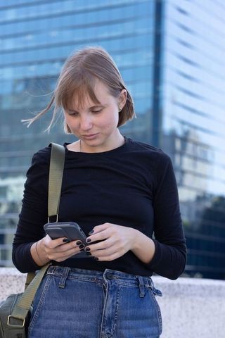Young woman checking phone while standing by ledge in urban plaza with glass skyscraper