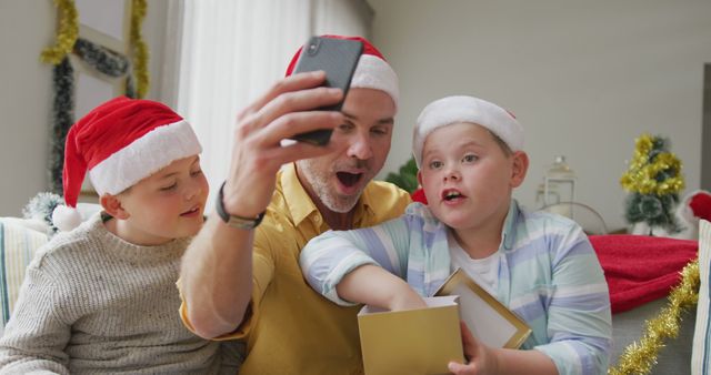 Caucasian father and two sons opening gifts while having a imagecall on smartphone during christmas. social distancing during covid 19 pandemic at christmas time