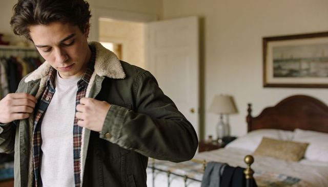 Young man adjusting shearling-collar jacket in cozy bedroom with brass bed and layers