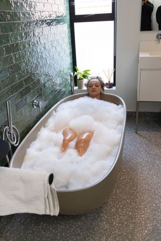 African american woman soaking in freestanding bubble bath by window, minimalist bathroom