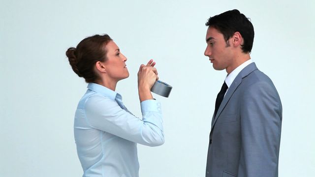 This video depicts a businesswoman attempting to place adhesive tape on a male colleague’s mouth against a plain white background. Both individuals are dressed in professional business attire, adding a humorous twist to a conventional office setting. Ideal for illustrating concepts of office humor, workplace dynamics, unconventional team interactions, and creative corporate communications.