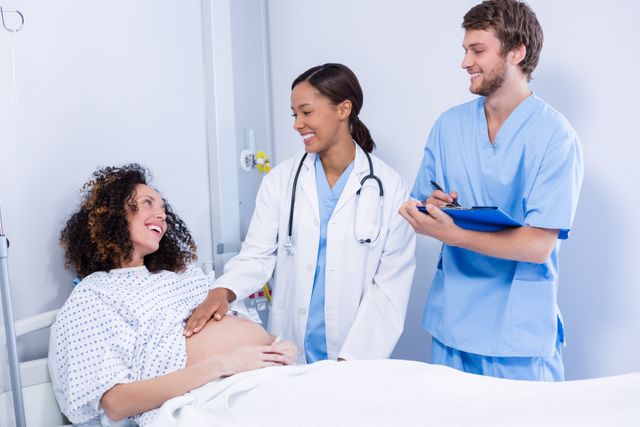 Medical professionals interacting with a pregnant woman in a hospital ward. The woman is smiling and appears to be in good spirits. This image can be used for healthcare, maternity care, prenatal care, and medical consultation themes. Ideal for websites, brochures, and articles related to pregnancy, healthcare services, and hospital environments.