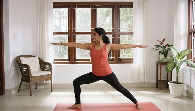 Indian woman practicing warrior ii yoga at home on pink mat by sunlit wooden windows, plants